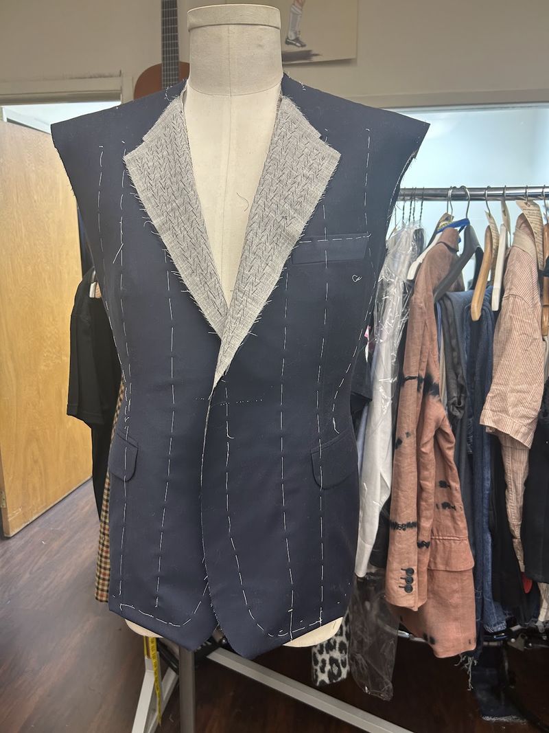 A navy pinstripe jacket in progress on a dress form, without sleeves, its lapels showing the inner canvas and white basting stitches
