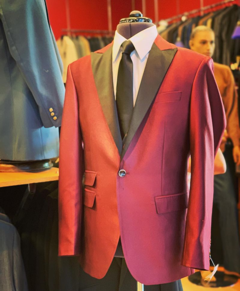 A red dinner jacket with black satin peak lapels on a mannequin in the shop, with racks of suits behind it