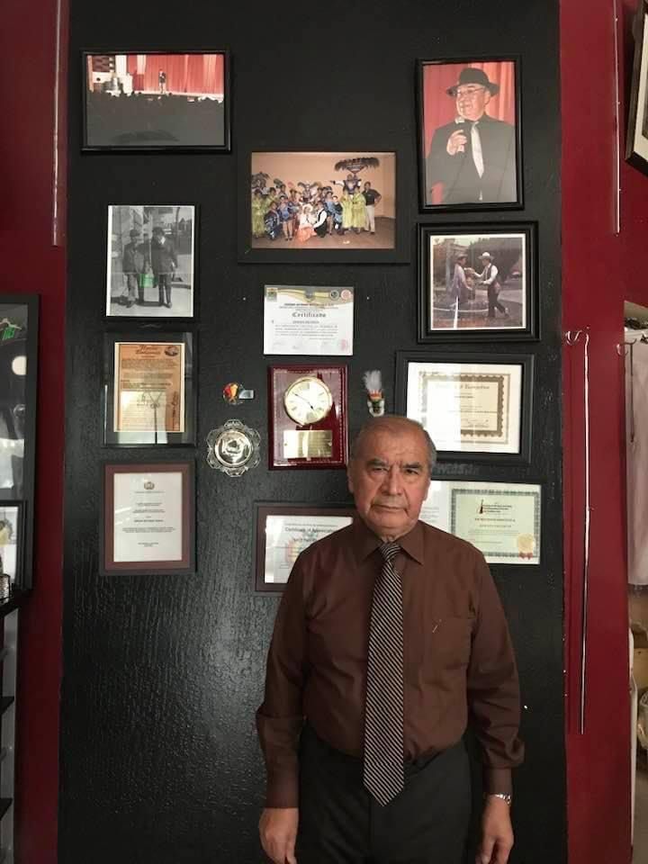 Efrain Escobar in a brown shirt and patterned tie, standing in front of a black wall of framed certificates, awards and photographs in his shop