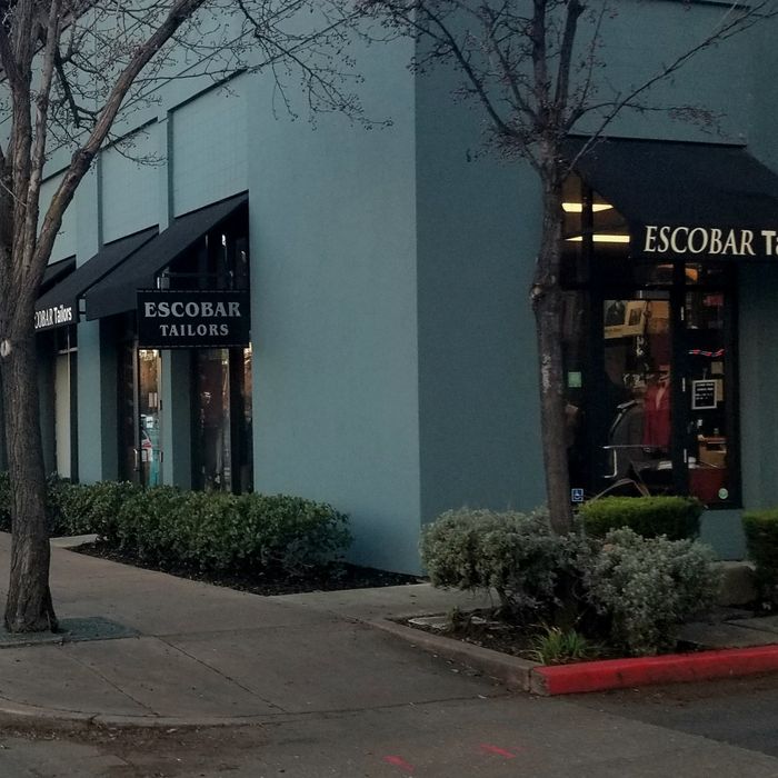 The shop's blue-gray corner storefront with black awnings reading Escobar Tailors, trees and planters along the sidewalk