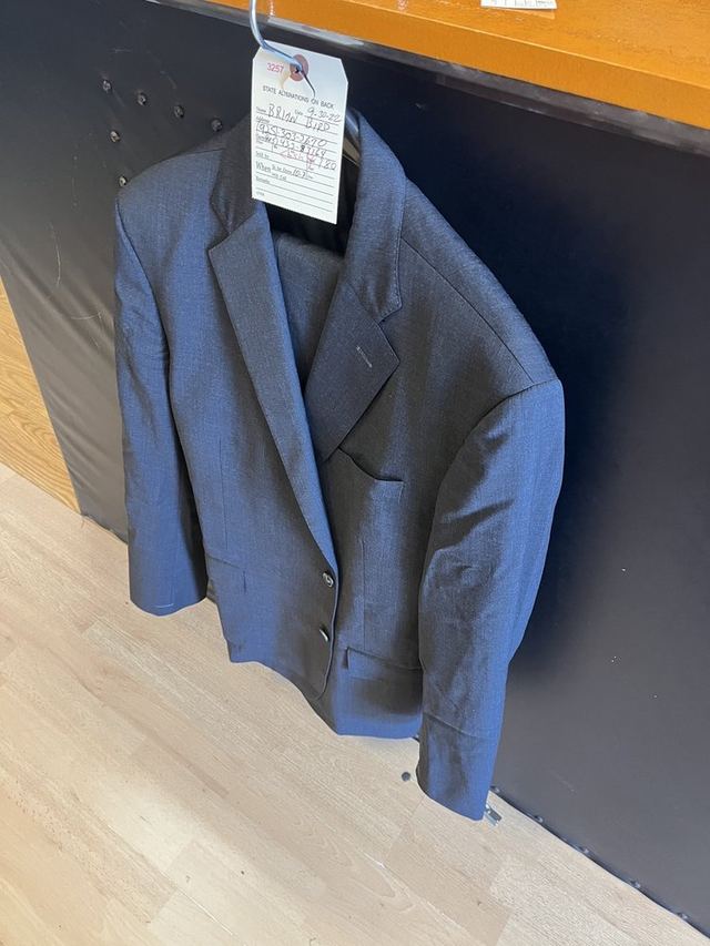 A blue sport coat on a hanger with a white paper ticket pinned to it, hanging against a dark panel by a wooden counter